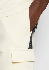 Cream cargo shorts made of soft fabric, featuring a side pocket with a black logo tag and a flat flap pocket on the leg.