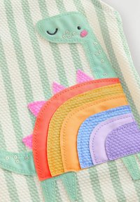 Der plüschige Teppich zeigt einen mintfarbenen Dinosaurier mit gestickten Details. Ein Regenbogen aus strukturierten, bunten Bögen in Rosa, Orange, Gelb und Lila.