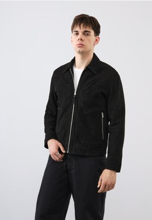 Giovane uomo che indossa una giacca in camoscio nera con zip sopra una maglietta bianca e pantaloni neri, in piedi davanti a uno sfondo chiaro e semplice.