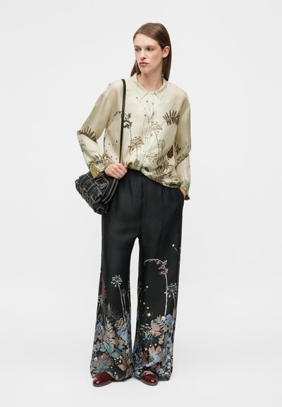 Blouse en soie à motif floral vert clair associée à un pantalon noir à jambes larges avec des imprimés botaniques colorés aux ourlets. Sac à épaule noir.