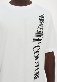 Balta medvilninė marškinėliai su vertikaliu juodu spaudiniu, kuriame parašyta „VERSACE JEANS COUTURE“, kairėje pusėje. Trumpomis rankovėmis, su apvaliu iškirpimu.