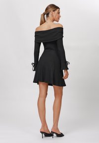 Abito nero a spalle scoperte in maglia con maniche a campana e dettagli con fiocchi. Lunghezza corta, tessuto leggero, texture liscia e design aderente.