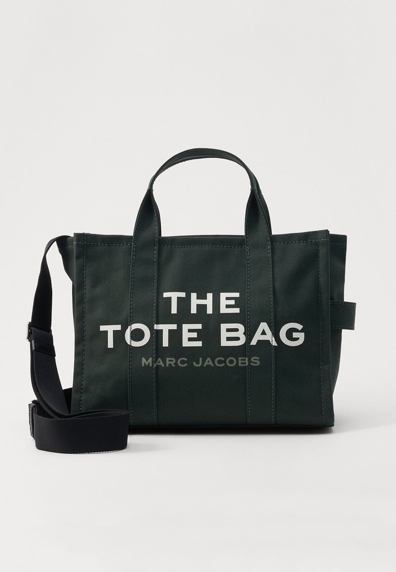 Tumši zaļš kanvas iepirkumu soma ar divām rokturiem, regulējamu melnu plecu jostu un tekstu "THE TOTE BAG MARC JACOBS" priekšpusē.