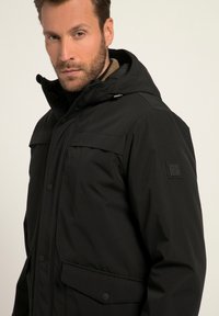 Giacca nera con cappuccio realizzata in tessuto morbido, con ampie tasche frontali, chiusura a zip e bottone, e un sottile dettaglio del logo sulla manica.