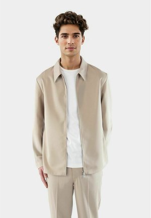 Beige zip-jakke med en glatt tekstur og krage, båret over en enkel hvit t-skjorte; parret med matchende beige bukser.