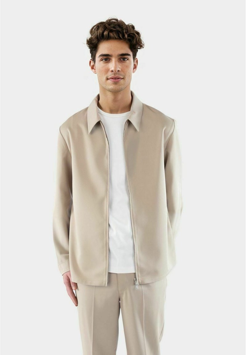 Beige zip-up jack met een gladde textuur en een kraag, gedragen over een eenvoudige witte t-shirt; gecombineerd met bijpassende beige broek.