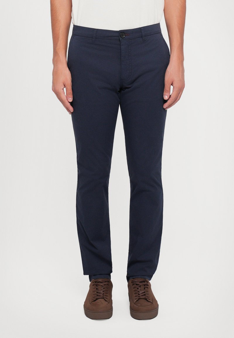Uomo che indossa pantaloni slim-fit blu navy con bottone frontale e zip, abbinati a scarpe stringate in suede marrone, in piedi davanti a uno sfondo bianco.