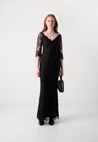 Robe de soirée en dentelle noire avec des manches trois-quarts, silhouette ajustée, décolleté en V et ourlet en forme de coquille. Accessoirisée avec un petit sac à main noir.