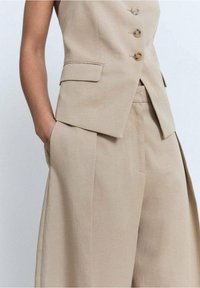 Beige op maat gemaakte gilet met vijf knopen, gecombineerd met brede broekspijpen. Voorzien van zakken en een schone, gestructureerde silhouette.