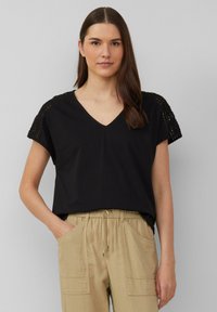 Haut noir à manches courtes avec un col en V et des détails en crochet aux épaules, associé à un pantalon beige avec cordon de serrage. Texture lisse.