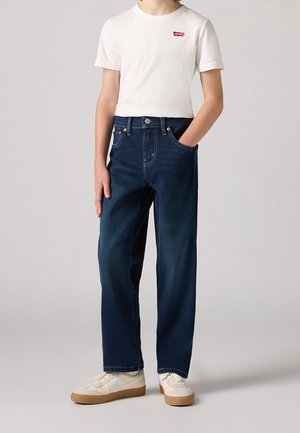 Persona con camiseta blanca de Levi's, vaqueros azul oscuro con la mano en el bolsillo y zapatillas blancas con suela de goma, de pie contra un fondo liso.