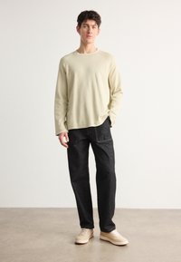 Light beige long-sleeve shirt with a crew neckline paired with dark denim trousers and light sneakers. Smooth texture, relaxed fit, minimal design.