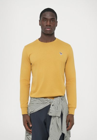 Tricou cu mâneci lungi, galben muștar, cu guler rotund și un mic logo brodat pe piept, purtat peste o cămașă în carouri legată în talie.