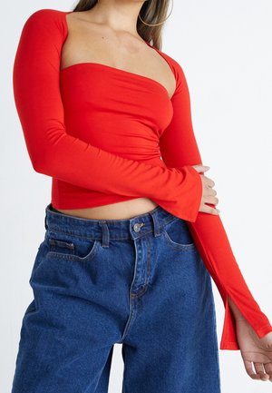 Rotes Langarmoberteil mit enganliegendem Oberteil, quadratischem Ausschnitt und ausgestellten Ärmeln, kombiniert mit hochtaillierten blauen Jeans.