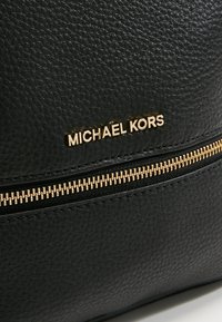 Gros plan sur du cuir noir texturé avec le logo Michael Kors doré et une fermeture éclair dorée sous le logo.