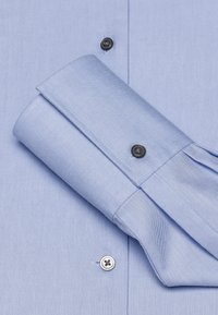 Camicia azzurro chiaro con colletto pieghettato, dotata di un singolo bottone nero e di un bottone bianco sul davanti, realizzata in tessuto liscio con una texture sottile.