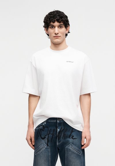 Νεαρός με σγουρά μαλλιά που φοράει λευκό T-shirt Off-White και σκούρα μπλε τζιν με μαύρα λογότυπα Off-White.