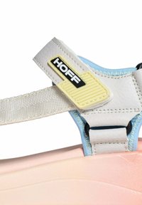 Sandalia blanca con una correa ancha, forro interior azul, logo amarillo y negro en la correa, y suela suave de color rosa. Detalles texturizados visibles.