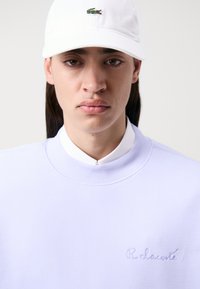 Gai violets krekls ar ribotu apkakli un izšūtu "R. Lacoste" detaļu, uzvilkts virs baltas apkakles krekla, apvienots ar baltu cepuri.