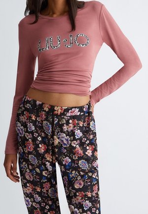 Kobieta ubrana w różowy top z długim rękawem z wzorzystym napisem "LIU·JO" oraz czarne spodnie z kolorowym kwiatowym wzorem.