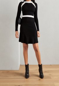 Vestido negro de canalé con mangas largas, cinturilla blanca y acentos de botones dorados en los hombros. Llevado con botas negras hasta el tobillo.