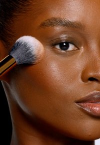 Make-up-Pinsel mit weichen, mehrfarbigen Borsten, der Produkt auf glatte, gebräunte Haut aufträgt. Der goldene Griff bildet einen Kontrast zur Textur des Pinsels.