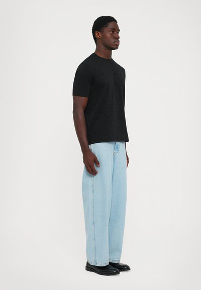 Mann, der seitlich steht und ein schwarzes Kurzarmhemd, hellblaue weite Jeans und schwarze Schuhe trägt, vor einem schlichten weißen Hintergrund.