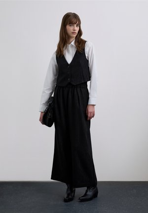 Gilet nero a righe fini indossato sopra una camicia bianca con bottoni, abbinato a pantaloni neri a gamba larga e stivaletti. Borsa nera completa l'outfit.