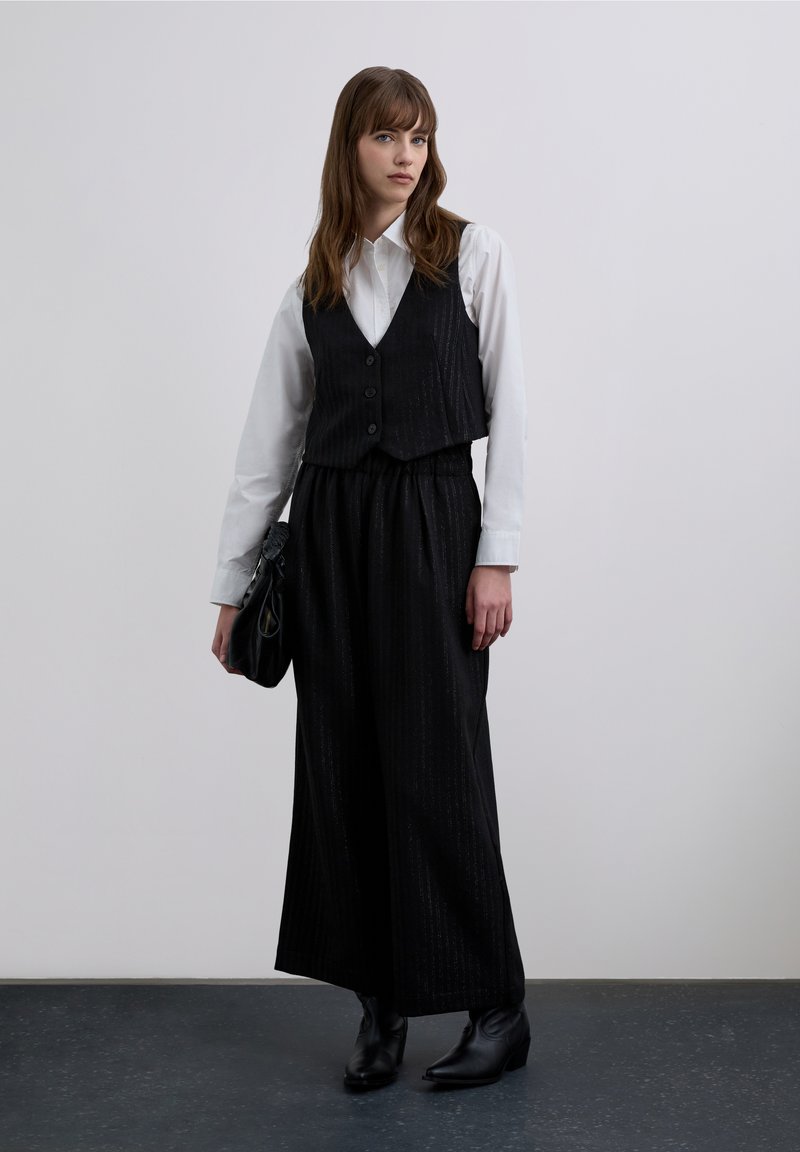 Czarna kamizelka w prążki noszona na białej koszuli zapinanej na guziki, zestawiona z czarnymi szerokimi spodniami i botkami za kostkę. Dopełnieniem stroju jest czarna torebka.