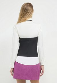 Corsetto nero con una texture liscia, indossato sopra una camicia bianca. Il modello indossa anche una gonna rosa a spina di pesce. I dettagli includono maniche lunghe e bottoni.