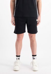 Zwarte katoenen shorts met een relaxte pasvorm, voorzien van een klein wit logo aan de zijkant. Gecombineerd met witte sneakers en zwart-witte sokken.