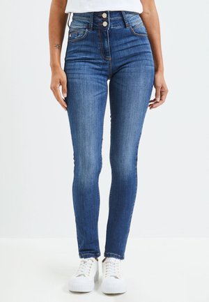 Femme portant un jean skinny bleu foncé taille haute avec fermeture à double bouton et baskets blanches, debout devant un fond uni.