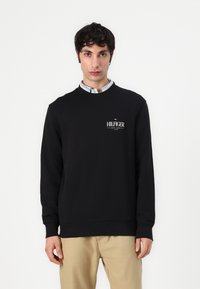 Juoda palaidinė su apvalia apykakle, ant kurios pavaizduotas baltas "HILFIGER NEW YORK" logotipas. Medžiaga atrodo lygi ir šiek tiek tampanti. Dėvima ant smėlio spalvos kelnių.