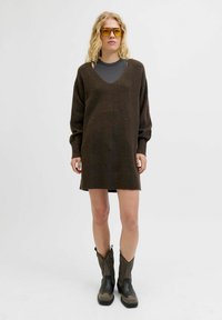 Vestido de punto marrón oversize con escote en V, mangas largas y puños acanalados, combinado con una camiseta gris de manga larga y botas de vaquero negras.