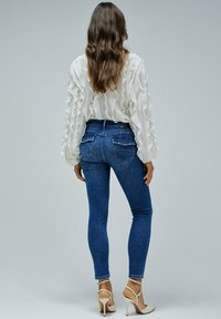 Blusa blanca con volantes y mangas largas, combinada con jeans ajustados de color azul oscuro que tienen bolsillos traseros y detalles de costura sutil. Tacones altos beige.