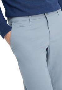 Pantaloni azzurri con una texture liscia, caratterizzati da una parte anteriore piatta, tasche laterali e un design slim fit. Abbinati a un maglione blu scuro.