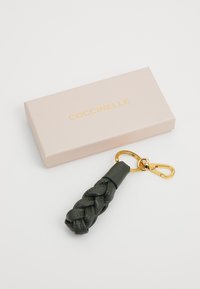 Geflochtenes dunkelgrünes Leder-Schlüsselanhänger mit einem goldenen Metallverschluss, neben einer hellrosa Box platziert, auf der "Coccinelle" in Gold gedruckt ist.
