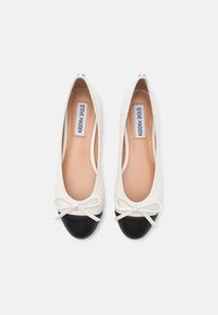 Ballerines avec une tige en simili cuir blanc et un bout en cuir verni noir, agrémentées d'un petit nœud sur le devant. Doublure intérieure lisse.
