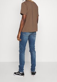 Persona de pie frente a una pared blanca, vistiendo una camiseta de rayas marrones, jeans azules y zapatillas Vans negras con suelas blancas.