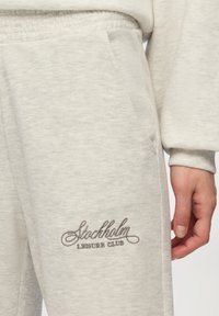 Jasnoszare dresy z miękkiej tkaniny, z boczną kieszenią i haftowanym logo "Stockholm Leisure Club" w ciemnoszarym kolorze.