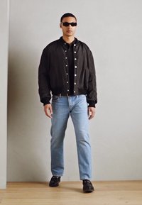 Veste bomber noire avec boutons pression, t-shirt noir, jeans bleu clair et chaussures noires. Le modèle porte des lunettes de soleil noires, debout sur un sol en bois.