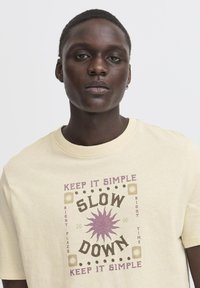 Lys gul bomull t-skjorte med et sentralt grafisk motiv med teksten "SLOW DOWN" i brun og lilla, omkranset av dekorative detaljer.