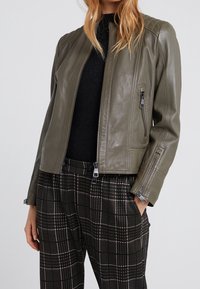 Oliivinvihreä nahkatakki, jossa on istuva muotoilu, vetoketjydetaljit ja tikatut yksityiskohdat hartioilla, yhdistettynä ruutukuosisisiin housuihin.