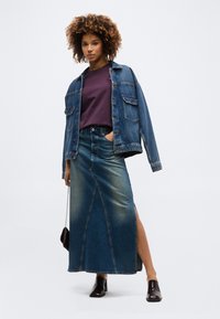 Γυναίκα με σγουρά μαλλιά που φοράει τζιν μπουφάν, μωβ πουκάμισο, μακρύ τζιν φούστα με πλαϊνή σχισμή, μαύρα μποτάκια μέχρι τον αστράγαλο, κρατώντας μια μικρή τσάντα.