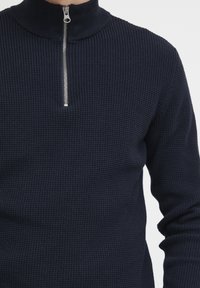 Pulover texturat, de culoare bleu marin, cu guler înalt și închidere cu fermoar pe jumătate; are un model în plasă și mâneci lungi.