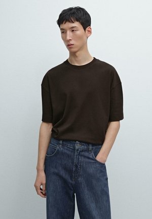 T-shirt marrone a maniche corte con scollo tondo, abbinata a jeans di denim blu. Il tessuto appare liscio, con una vestibilità comoda e dettagli minimi.