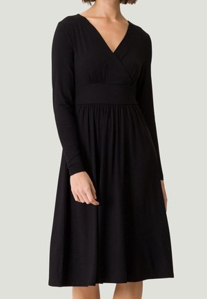 Mujer con un vestido midi negro de manga larga, escote en V y cintura fruncida, de pie frente a un fondo claro y liso.