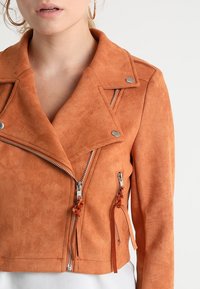 Lyhyt oranssi mokkanahkainen moottoripyörätakki, jossa on kauluskäänne, hopeiset metalliosat, kaksi vetoketjullista taskua ja koristeelliset nauhat vetoketjuissa.