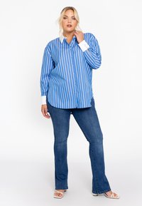 Blauw-wit gestreepte button-up shirt met een witte kraag, gecombineerd met uitlopende blauwe jeans en witte sandalen. Soepele stof, lange mouwen.