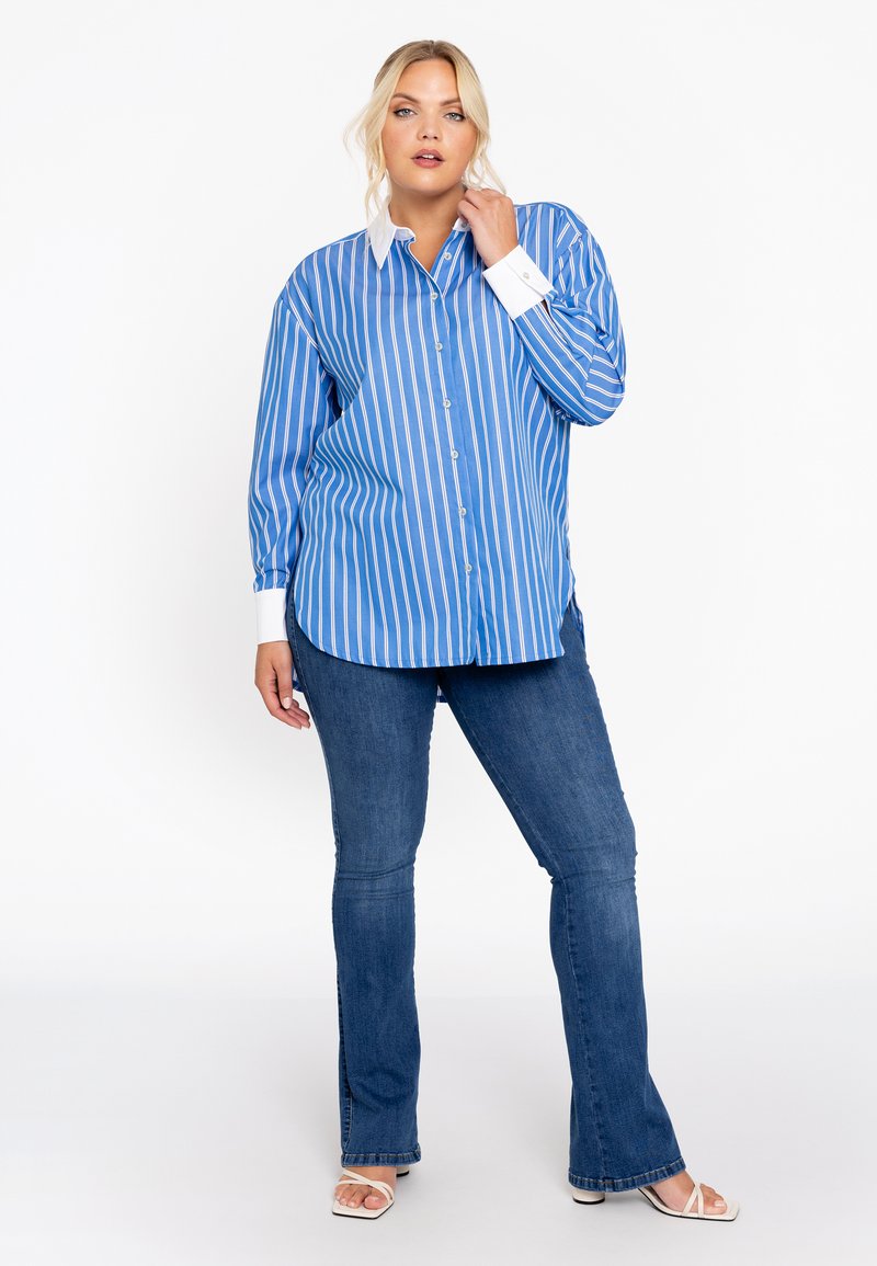 Blauw-wit gestreepte button-up shirt met een witte kraag, gecombineerd met uitlopende blauwe jeans en witte sandalen. Soepele stof, lange mouwen.